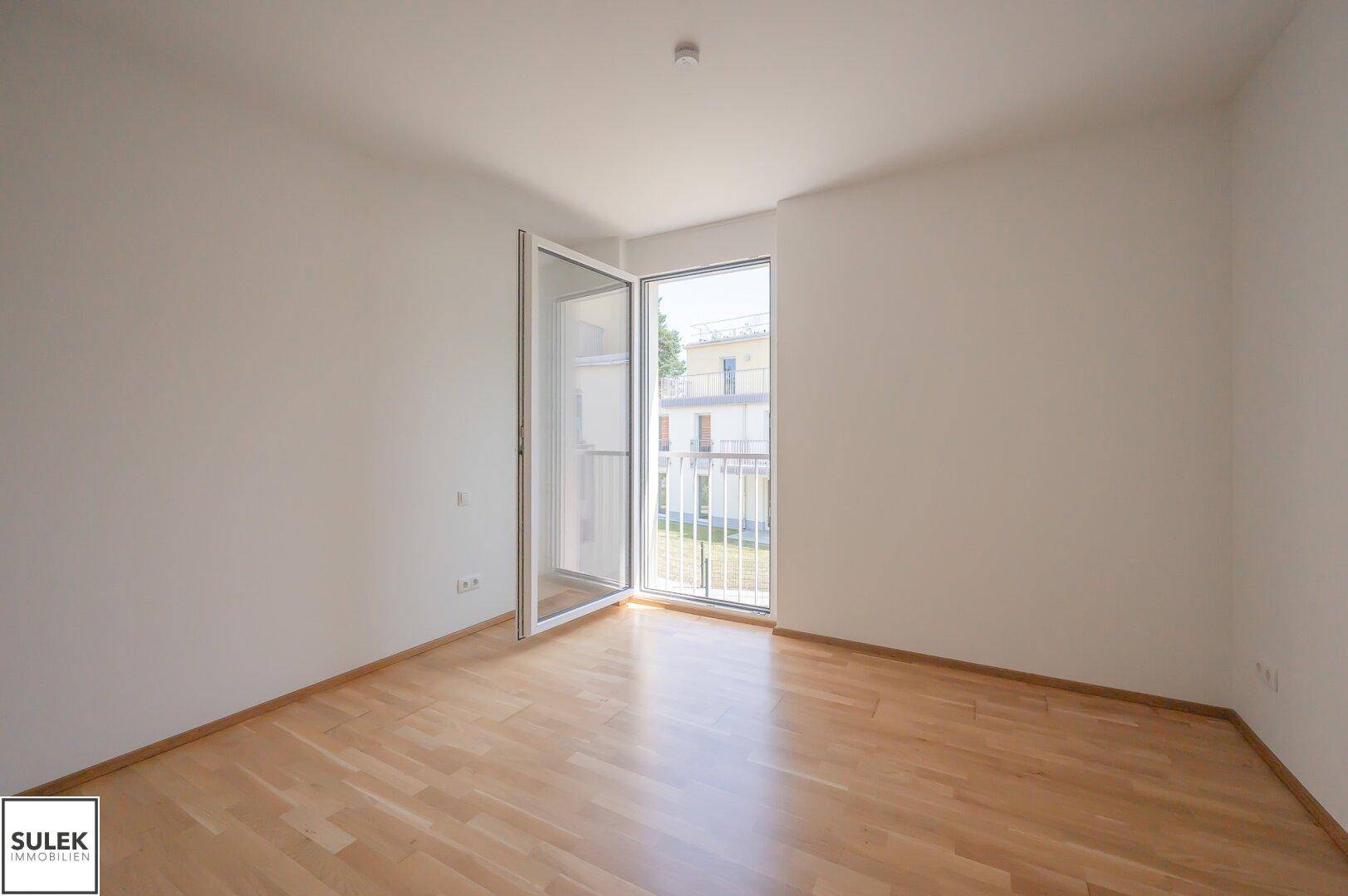 Leeres Schlafzimmer mit hellem Parkettboden und großem Fenster, das zu einem französischen Balkon führt.
