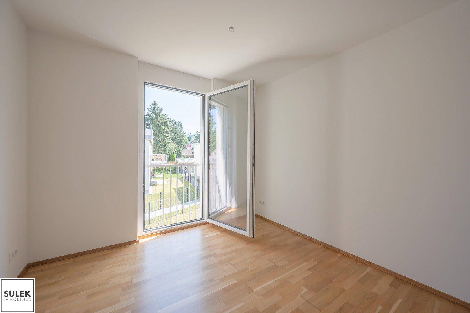 Helles Zimmer mit Parkettboden und Zugang zu einem französischen Balkon mit Blick ins Grüne.