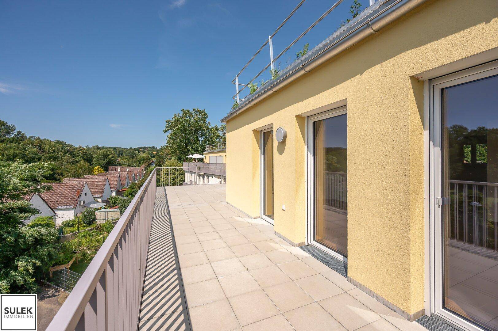 Langer Balkon mit Fliesenboden und Blick auf die umliegende Wohngegend und grüne Landschaft.