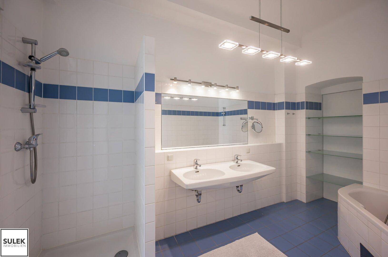 Badezimmer mit Duschbereich, Doppelwaschbecken und weiß-blauen Wand- und Bodenfliesen.