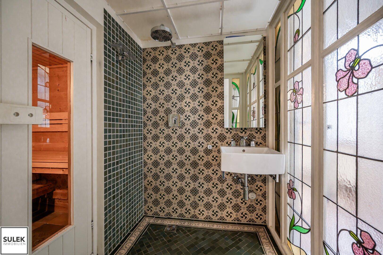 Badezimmer mit Dusche, Waschbecken, gemusterten Fliesen und Zugang zu einer Sauna.