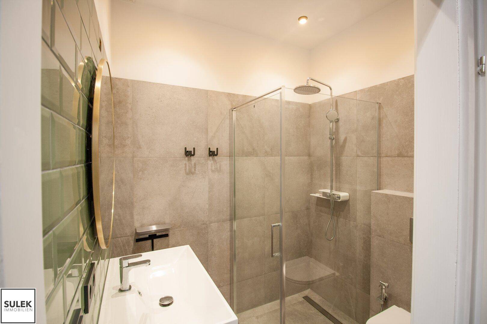 Modernes Badezimmer mit begehbarer Dusche, hellgrünen Fliesen und einem runden Spiegel.