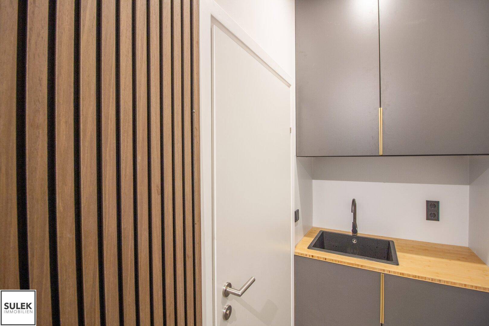 Kleine Küchenzeile mit dunklen Schränken, Holztheke und Spülbecken neben einer Holzwand.