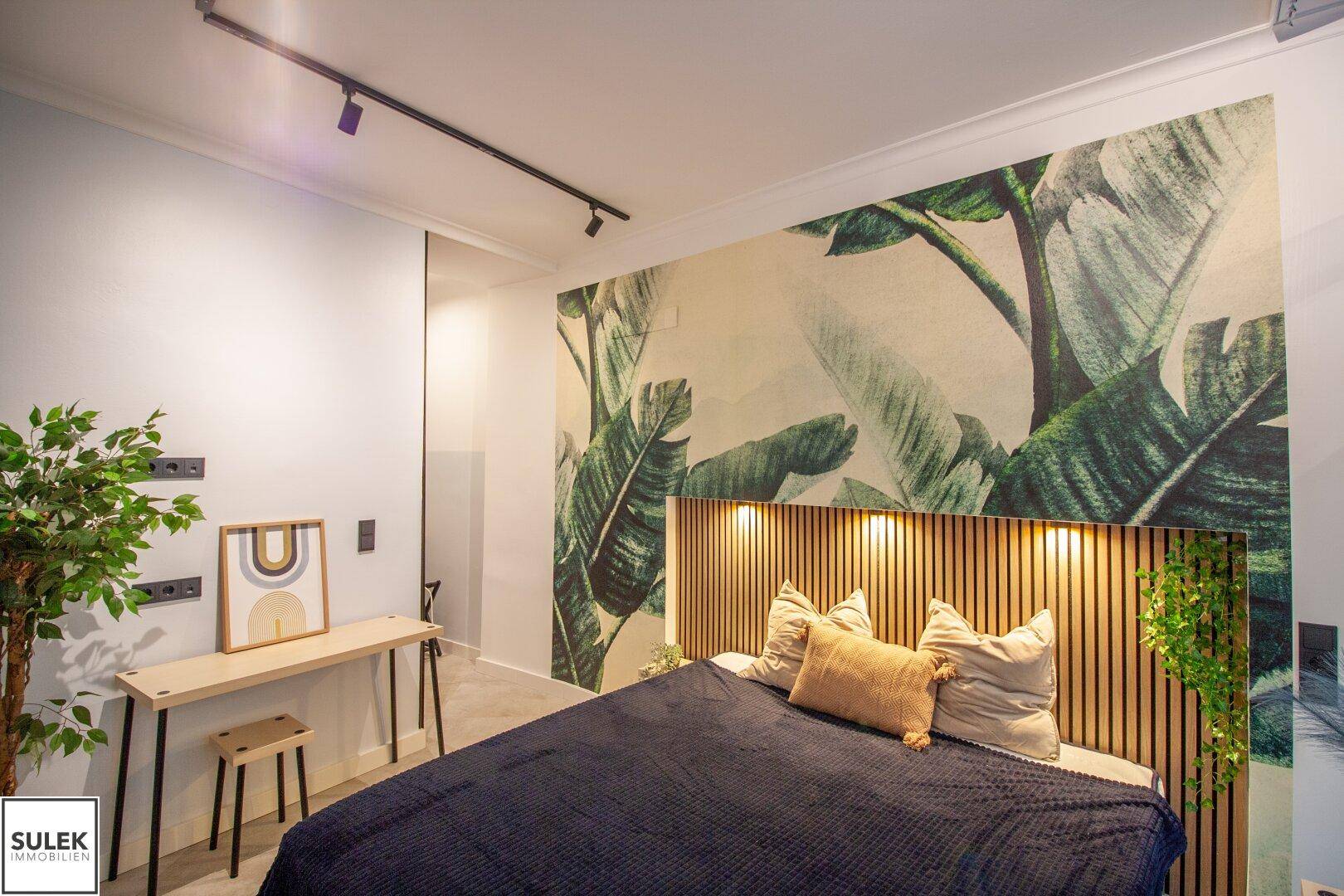 Schlafzimmer mit Holzkopfteil, tropischer Wandtapete und modernem Schreibtisch, gut beleuchtet.