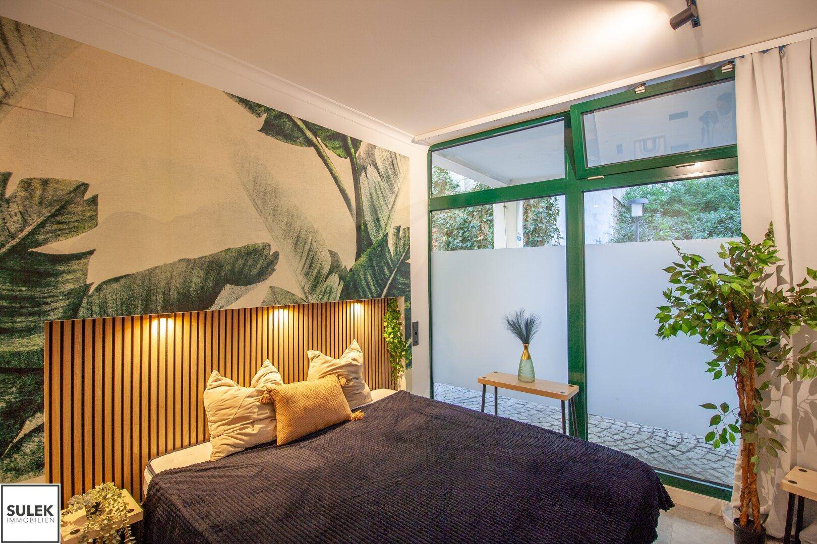 Schlafzimmer mit großem Bett, Holzkopfteil und tropischer Wandtapete, bietet Blick auf den Innenhof.