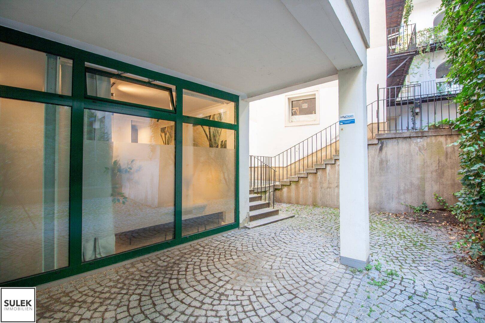 Gepflasterter Innenhof mit Zugang zu einem Gebäude mit großen Fenstern und einer Treppe.