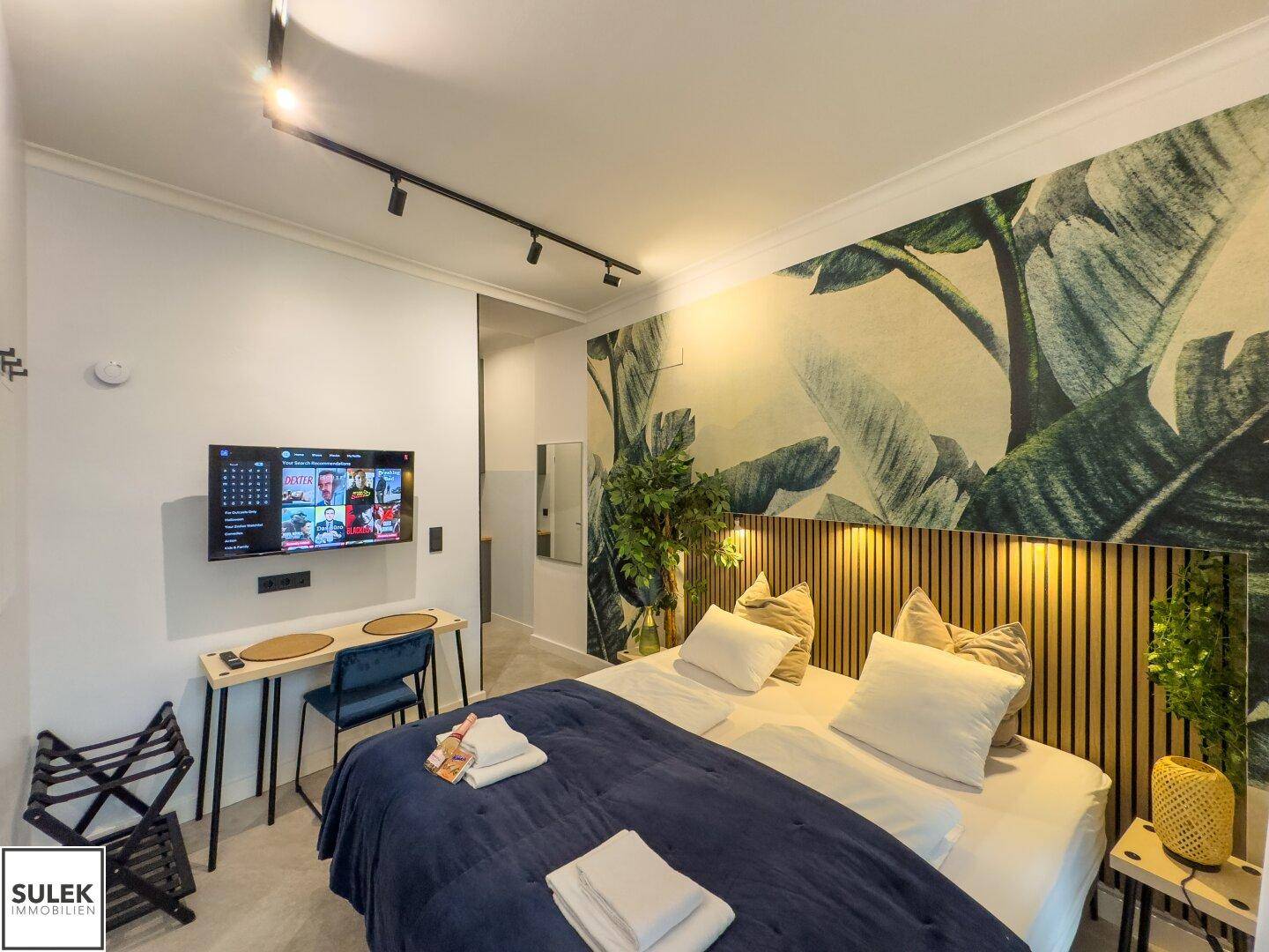 Schlafzimmer mit Wandfernseher, Holzkopfteil und tropischer Wandtapete, modern eingerichtet.