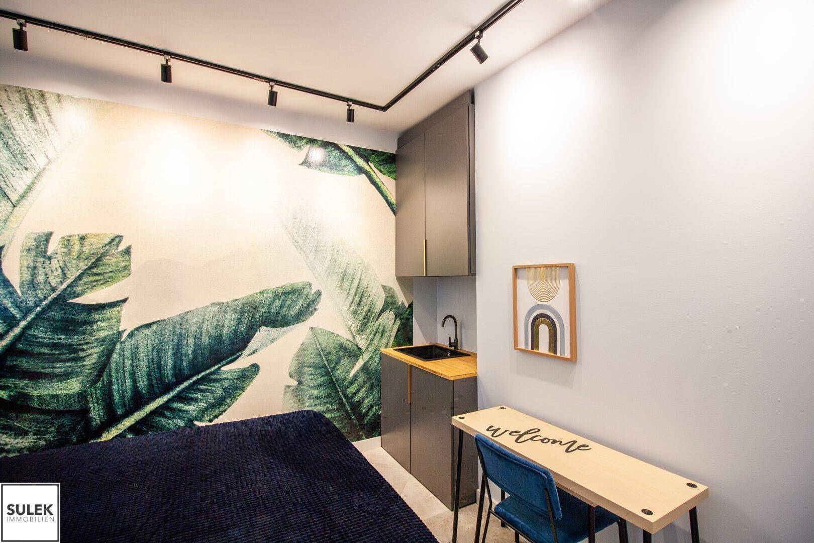 Schlafzimmer mit integrierter Küchenzeile, Schreibtisch und tropischer Wandtapete, modern gestaltet.