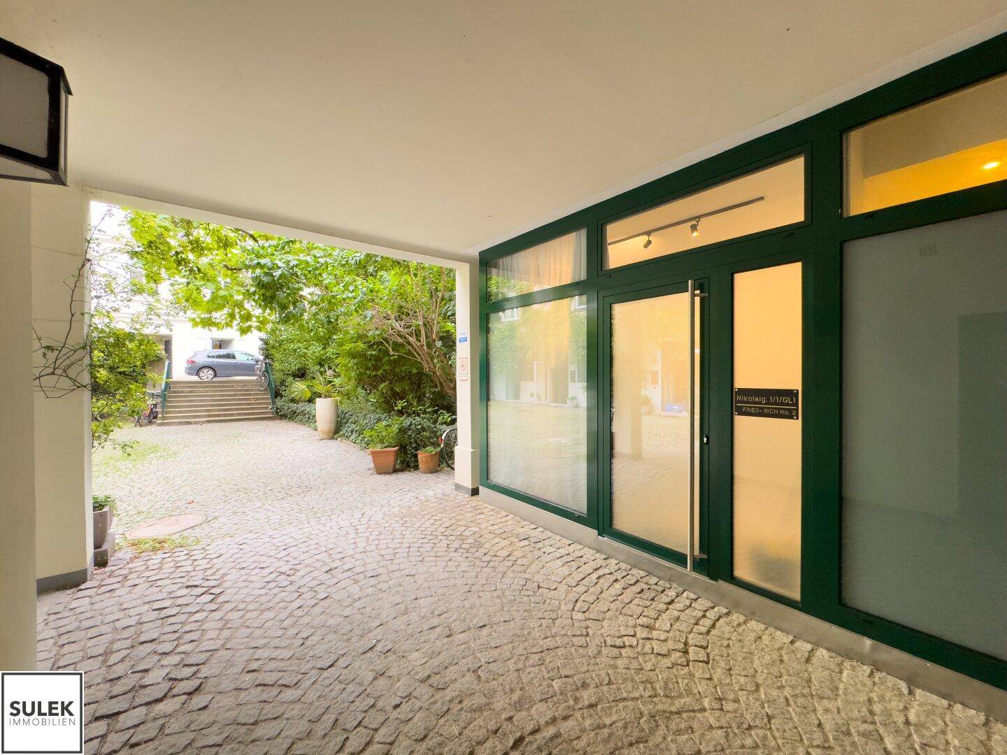 Gepflasterter Innenhof mit Zugang zu einem Gebäude mit großen Glasfenstern und Grünflächen.