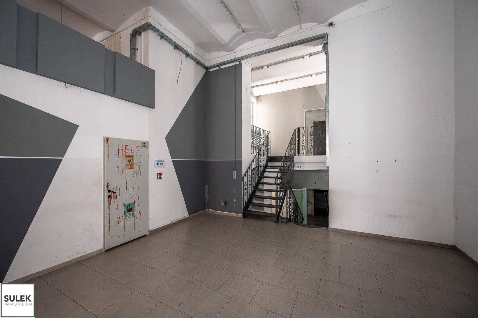 Großer Innenraum mit Fliesenboden, geometrischen Wandmalereien und einer schmiedeeisernen Treppe.