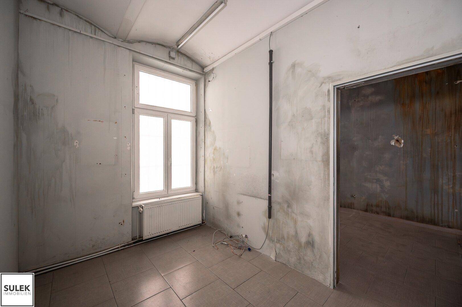Kleiner Raum mit Fliesenboden, Heizkörper unter dem Fenster und deutlichen Gebrauchsspuren an den Wänden.