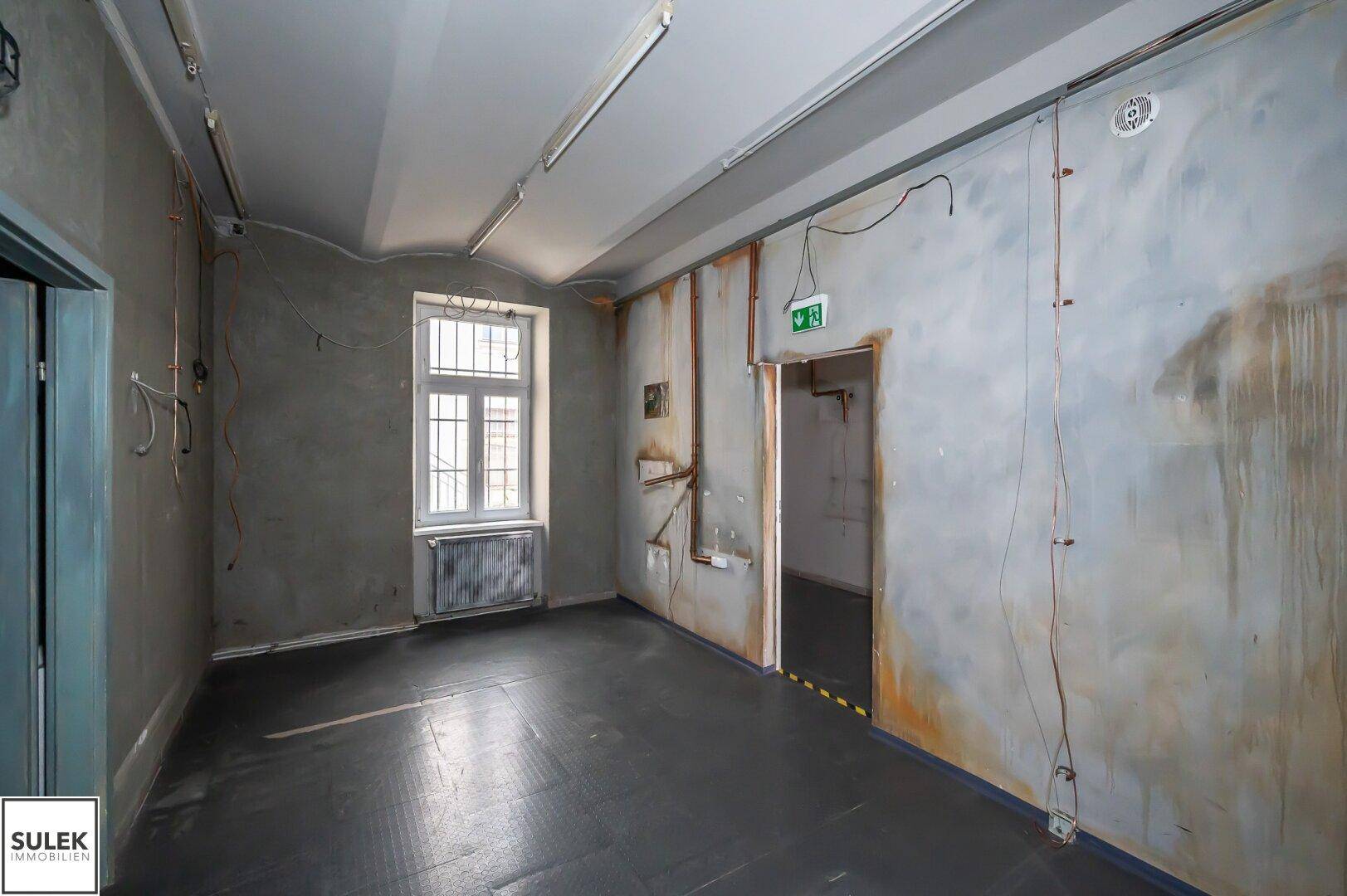 Raum mit dunklem Boden, sichtbaren Leitungen und stark renovierungsbedürftigen Wänden.