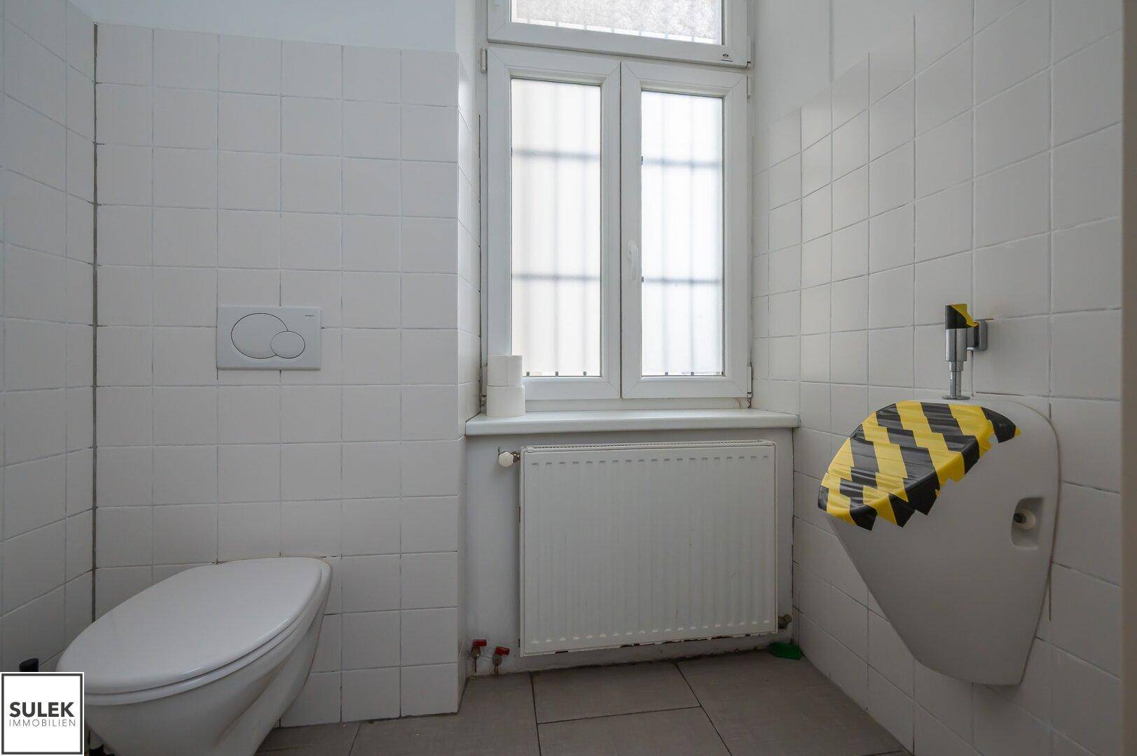 Badezimmer mit Toilette, Urinal, Fenster und weißen Fliesen, das eine Renovierung benötigt.