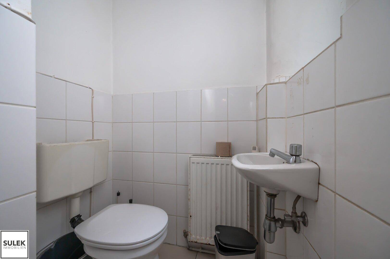 Einfaches Badezimmer mit Toilette, kleinem Waschbecken und weißen Fliesen an den Wänden.