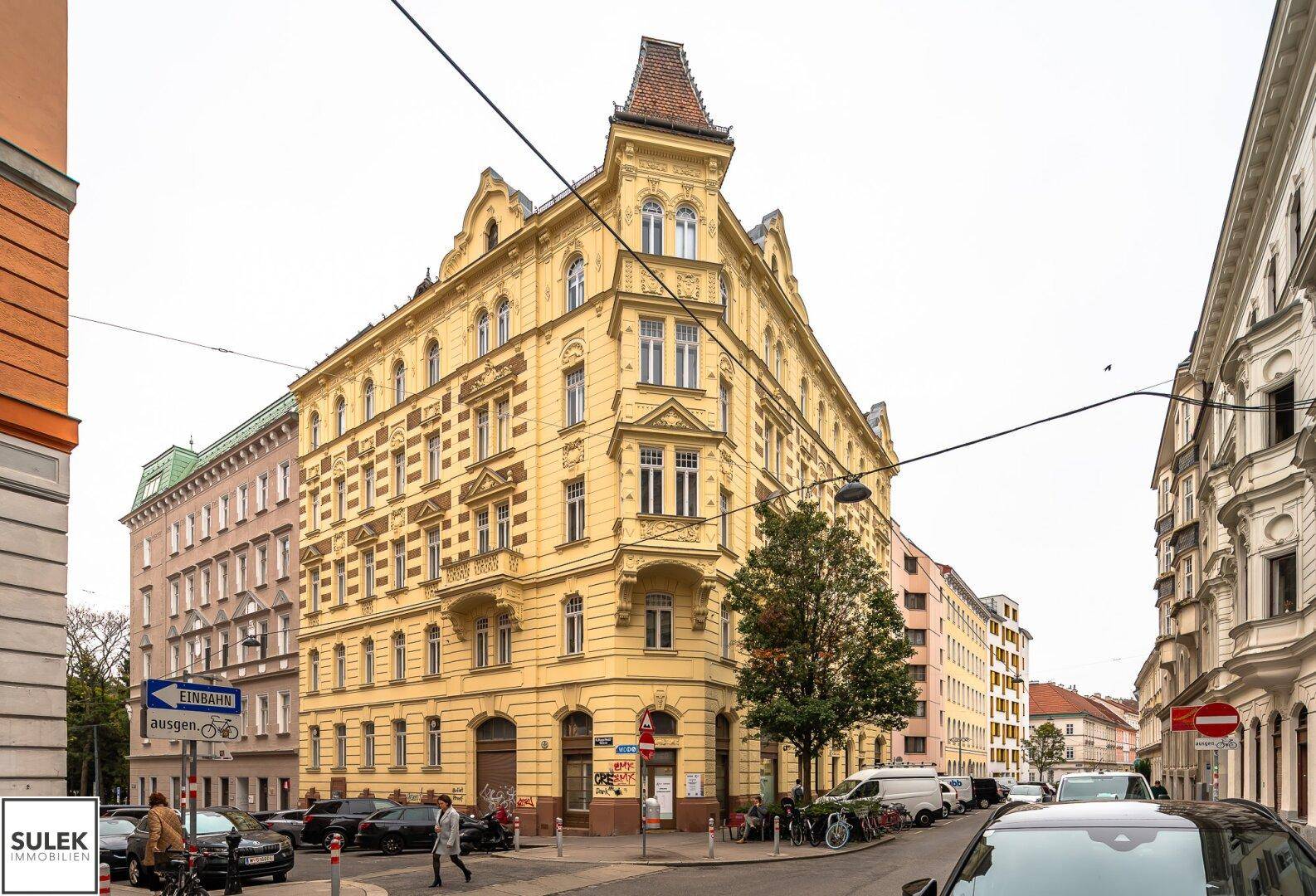 Historisches Eckgebäude mit gelber Fassade, vielen Fenstern und einem markanten Turmdach in der Stadt.