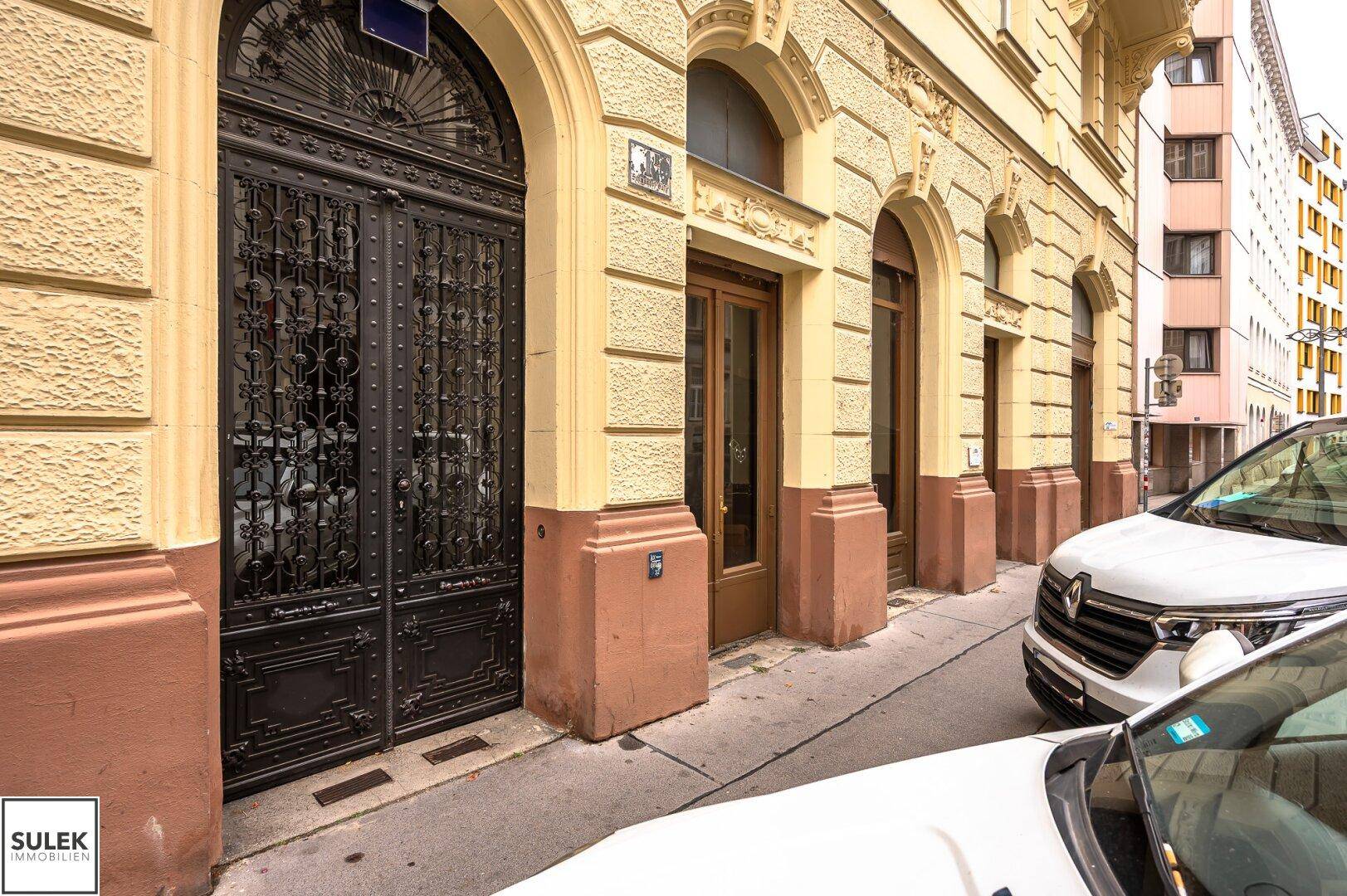 Historische Fassade eines Gebäudes mit kunstvollen Eingangstüren und geparkten Autos auf der Straße.
