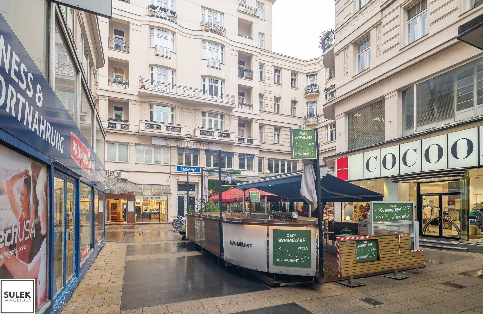 Lebhafte Einkaufsstraße mit Geschäften, Cafés und Wohngebäuden im Hintergrund.