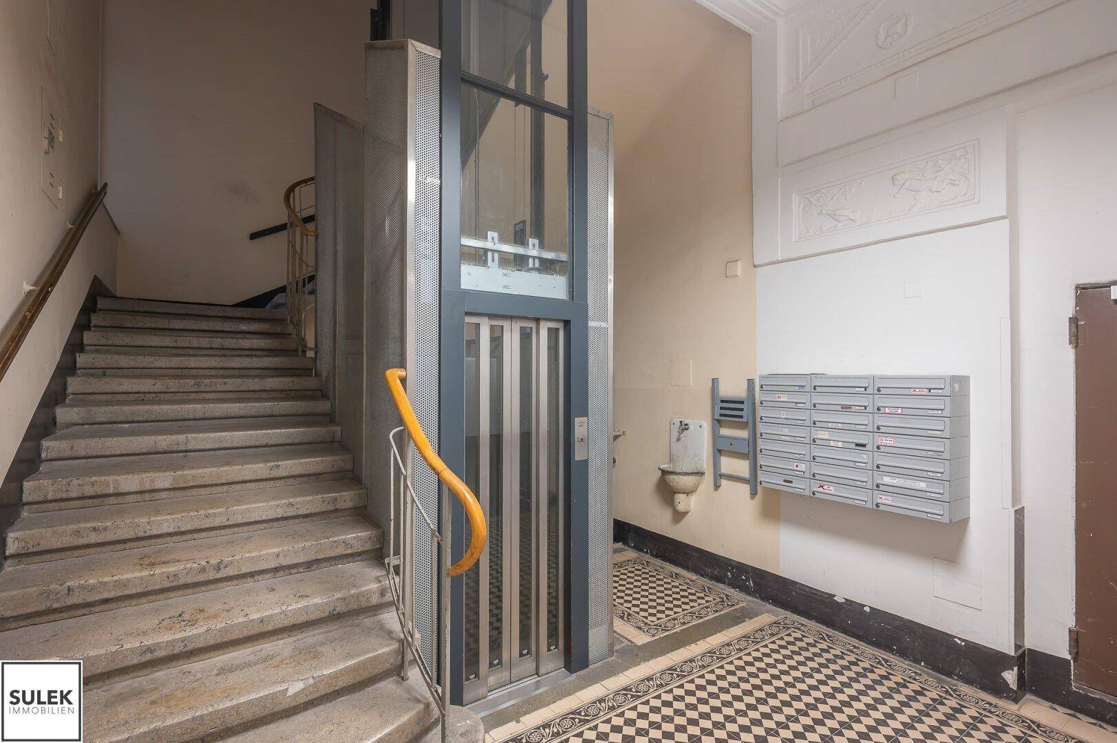 Eingangsbereich eines Altbaus mit modernem Glasaufzug, Steintreppe und historischen Bodenfliesen.