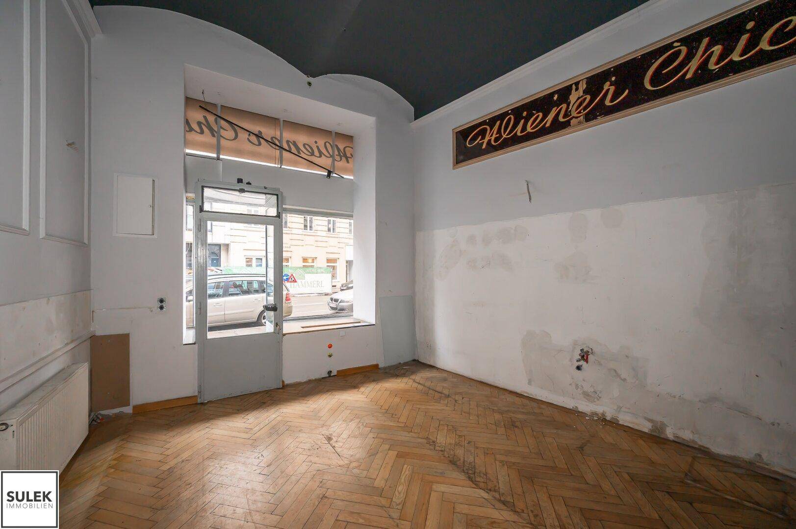 Gewerberaum mit großem Schaufenster zur Straße, Fischgrätparkett und renovierungsbedürftigen Wänden.