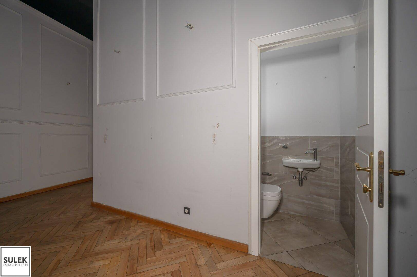 Gewerberaum mit Fischgrätparkett und Blick in eine moderne Gästetoilette mit Wand-WC und Waschbecken.
