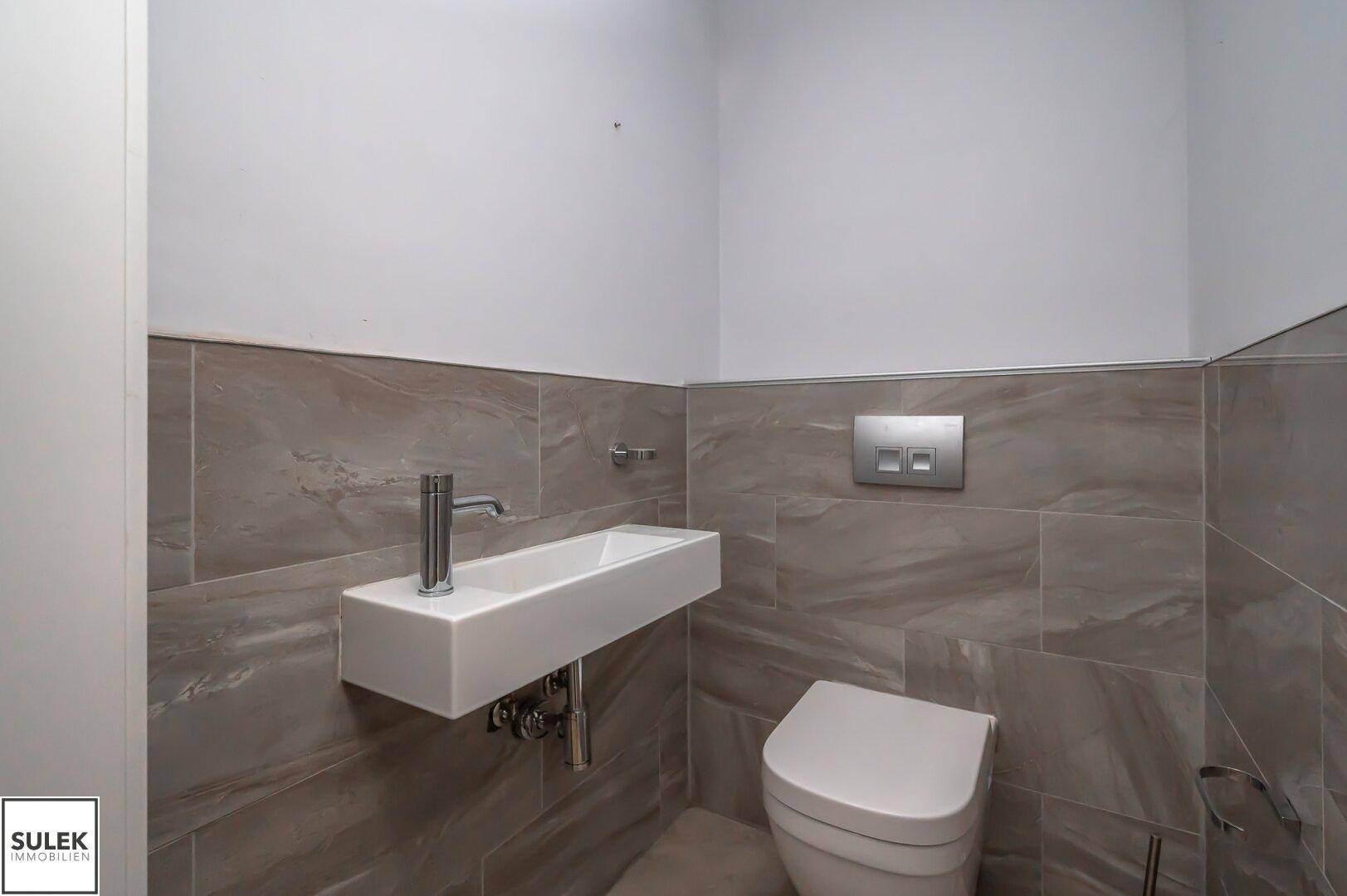Moderne Gästetoilette mit Wand-WC, kleinem Waschbecken und grauen Fliesen an den Wänden.