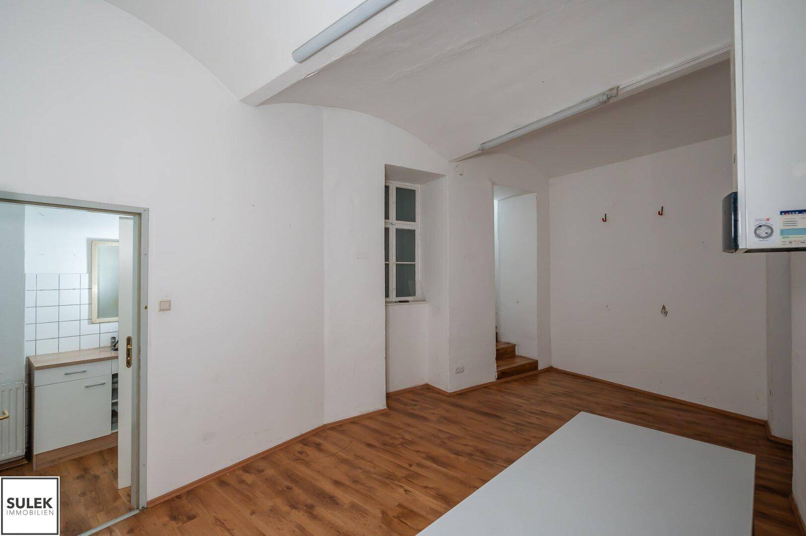 Gewerberaum mit Holzfußboden und Blick in eine kleine Küchenzeile mit Spülbecken und Arbeitsplatte.