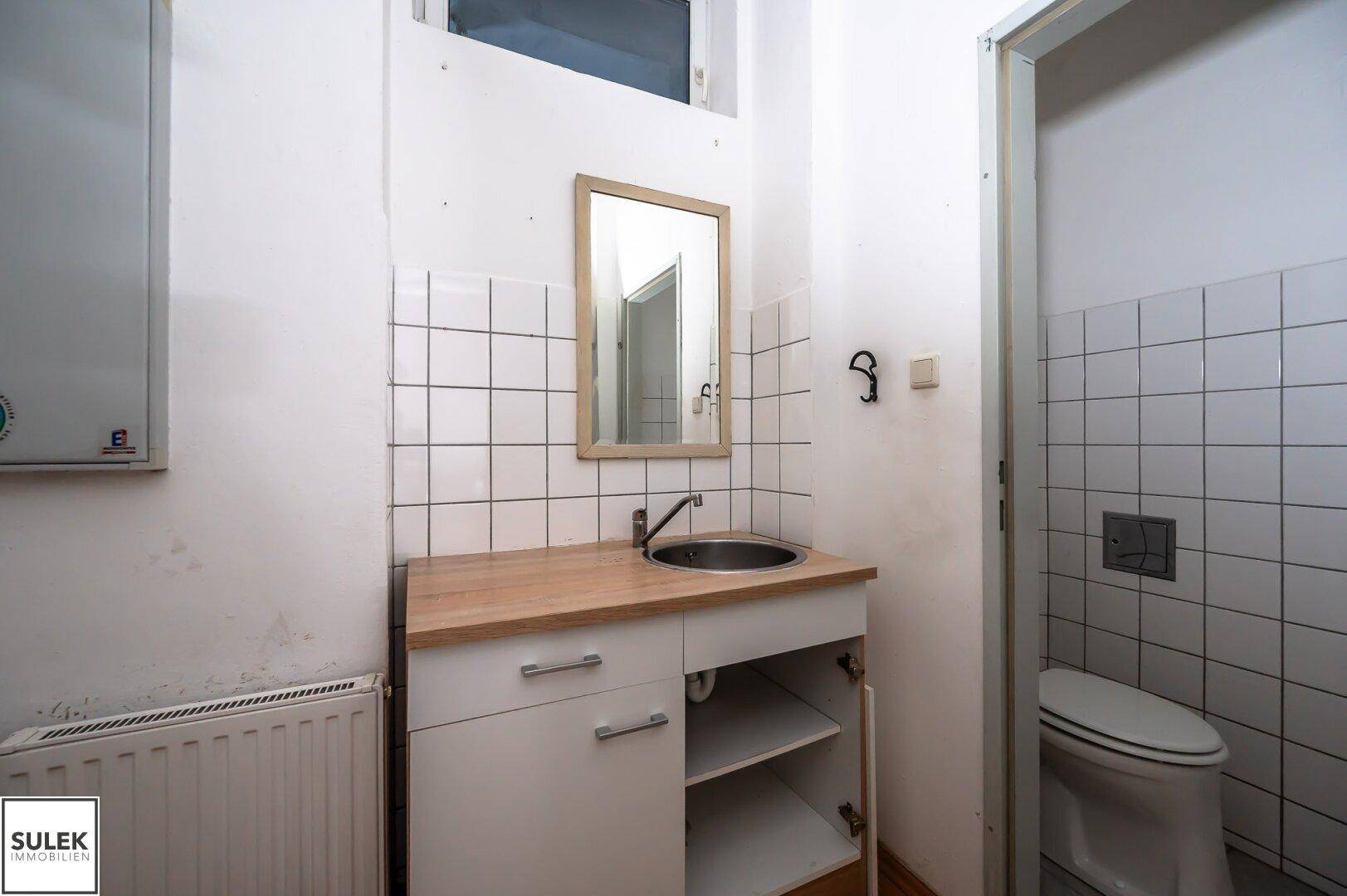Kleine Küchenzeile mit Spülbecken, Arbeitsplatte und angrenzender Toilette, umgeben von weißen Fliesen.