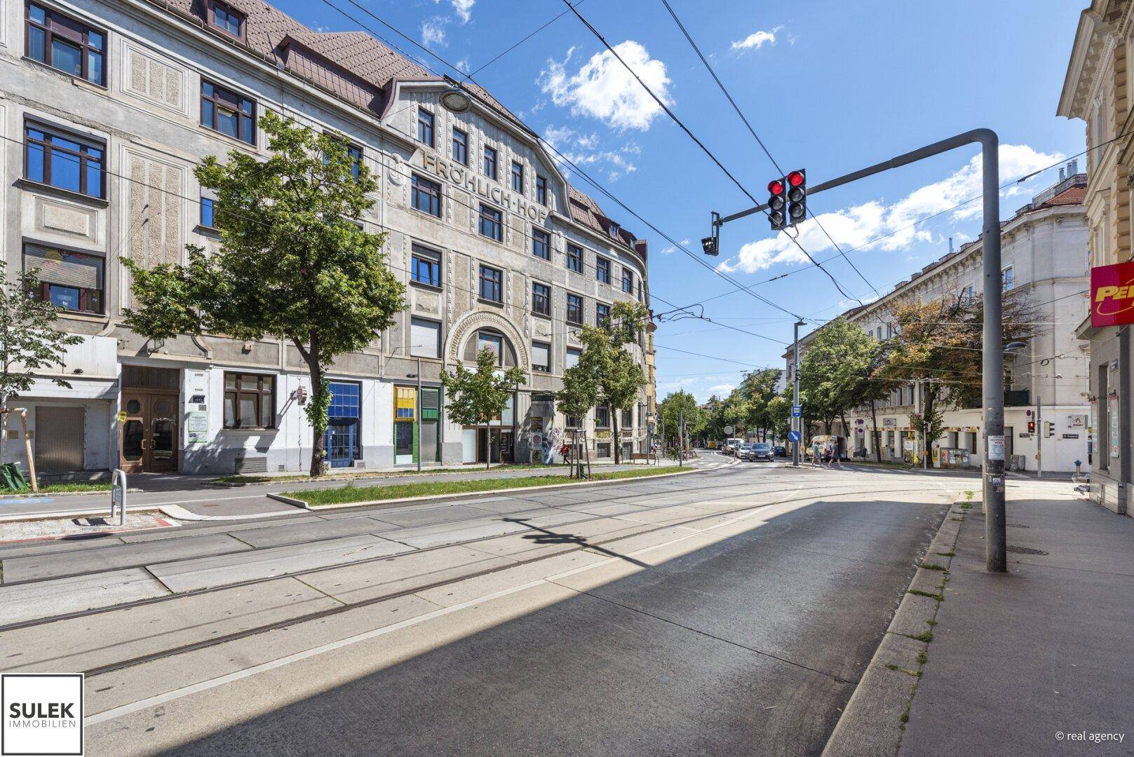 Weitläufige Straßenansicht mit dem historischen Gebäude "Fröhlich-Hof" und einer Straßenbahnhaltestelle.
