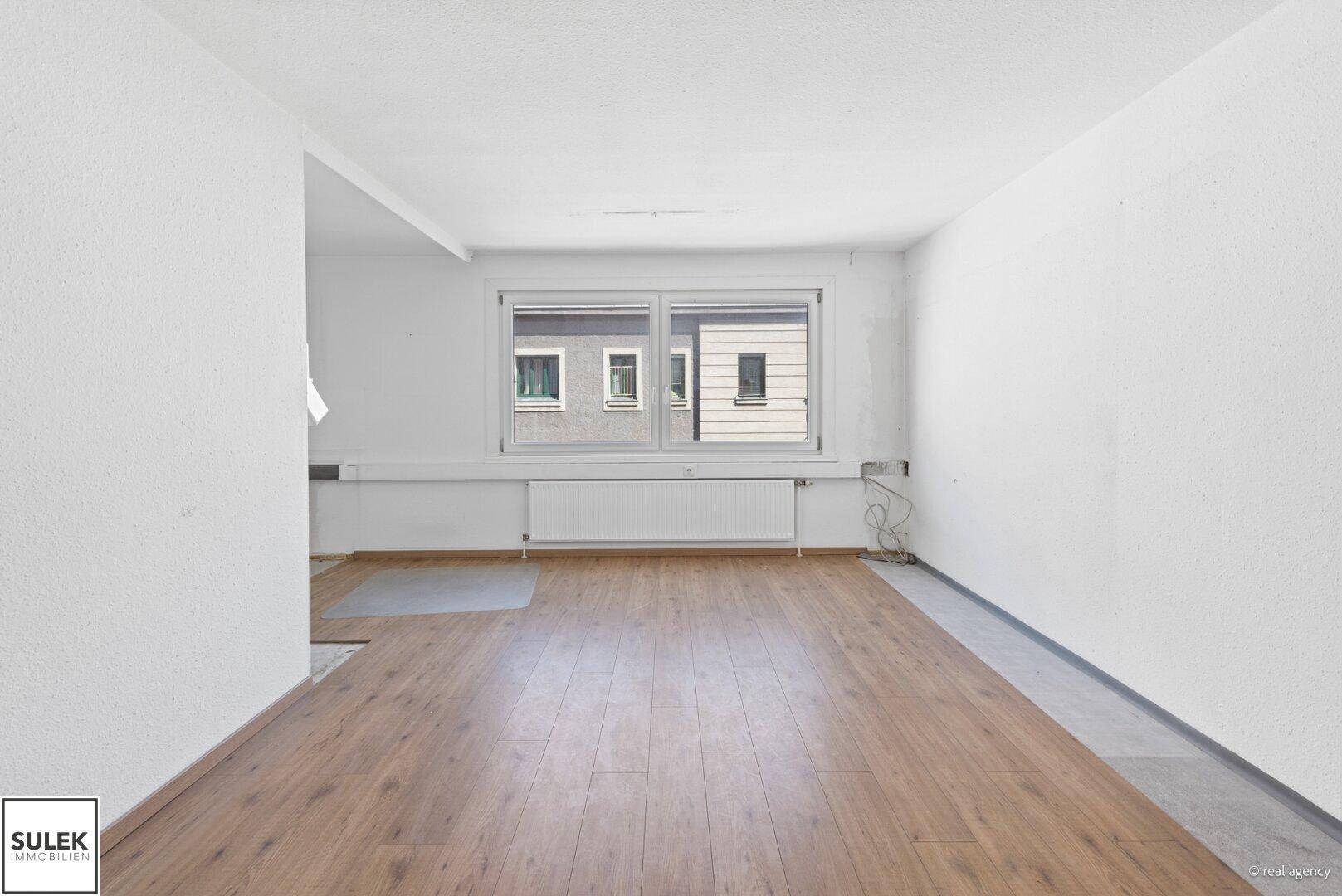 Leerer Raum mit Holzfußboden und einem großen Fenster, das viel Tageslicht hereinlässt.