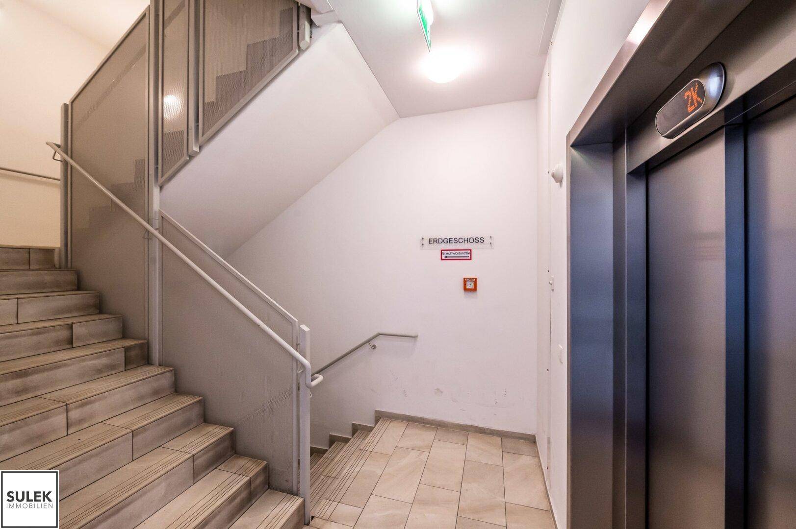 Treppenhaus und Aufzug in einem Mehrfamilienhaus mit Hinweisschildern.