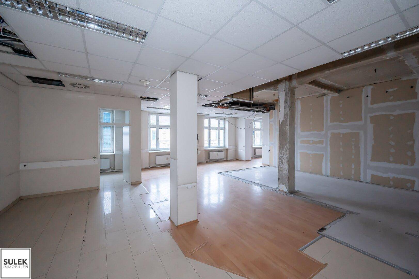 Großer Büroraum mit gemischten Bodenbelägen und sichtbaren Renovierungsarbeiten an Wänden und Decke.