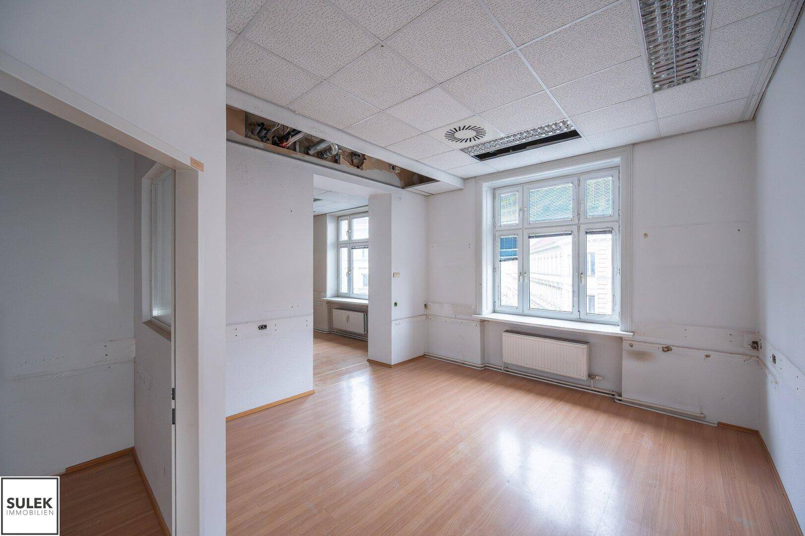 Heller Büroraum mit großen Fenstern, Laminatboden und sichtbaren Heizkörpern, der renoviert werden muss.