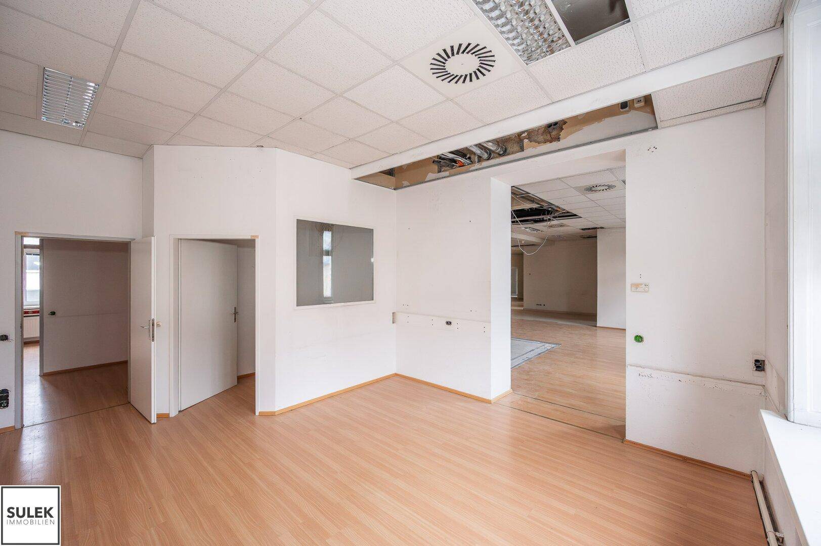 Geräumiger Büroraum mit Laminatboden und abgehängter Decke, der Renovierungsbedarf aufweist.