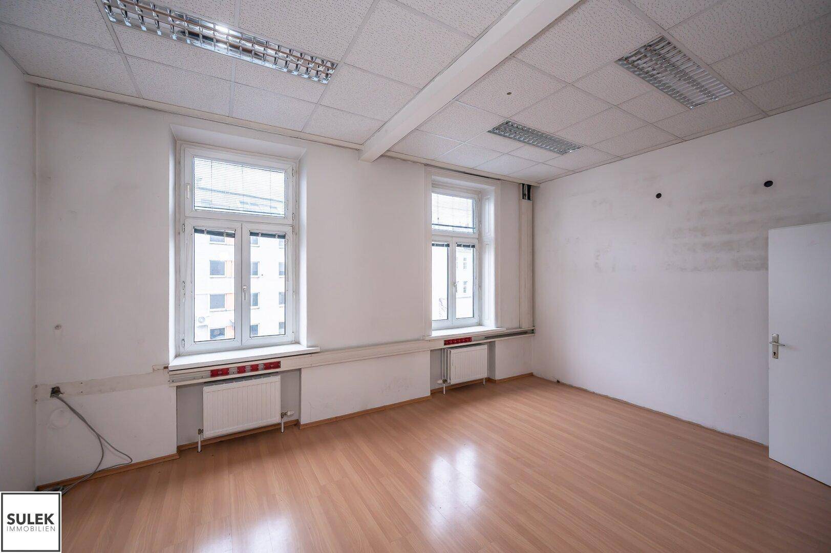 Helles Einzelbüro mit zwei großen Fenstern, Laminatboden und Heizkörpern unter den Fenstern.