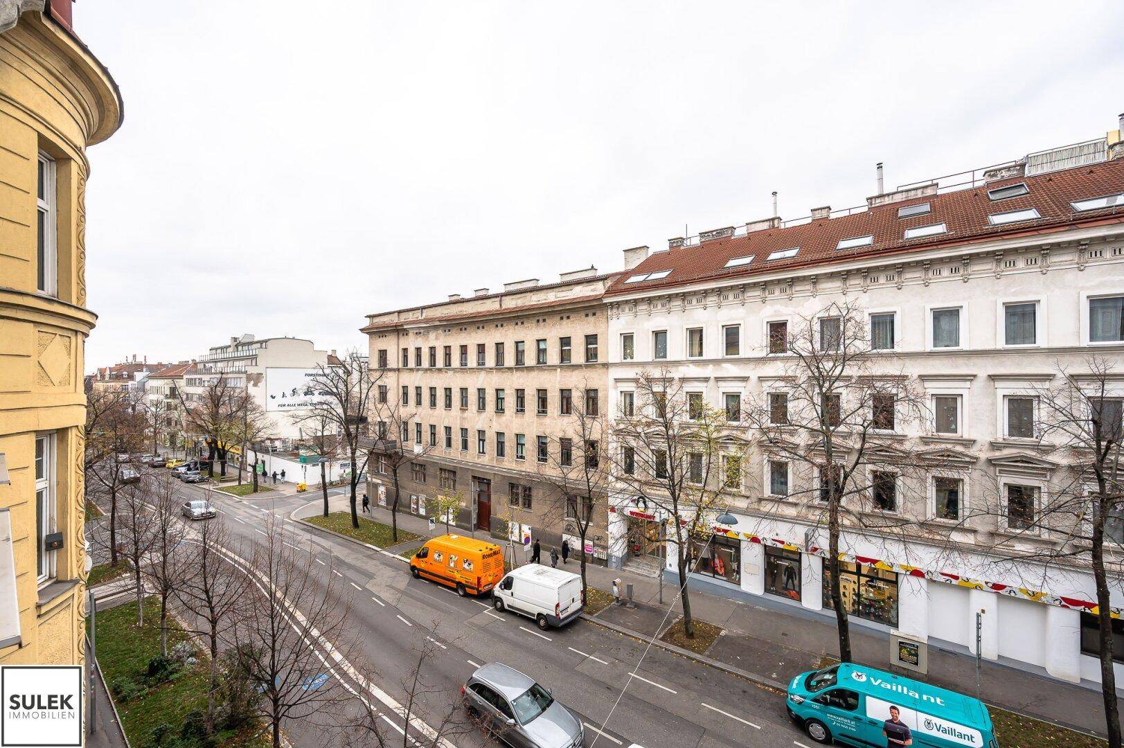 Weitläufiger Blick auf eine Stadtstraße mit Gebäuden und Verkehr, unter einem bewölkten Himmel.