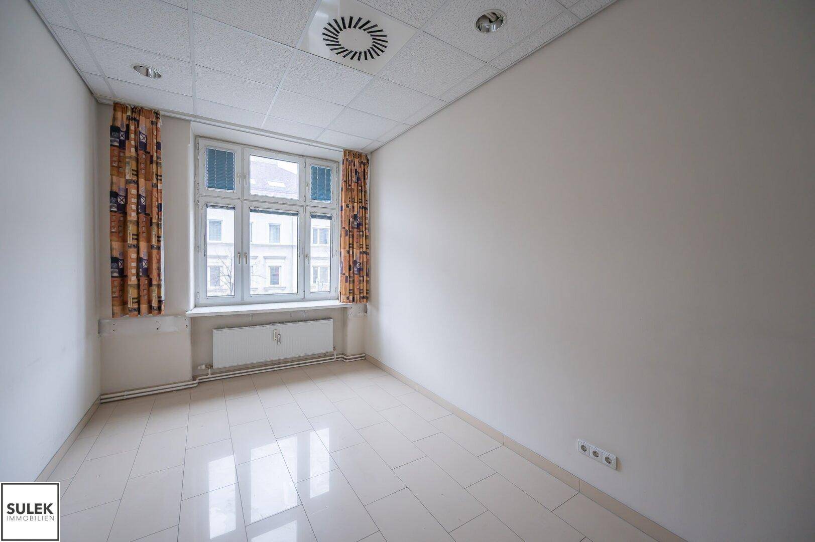 Helles Einzelbüro mit Fliesenboden, einem Fenster und Vorhängen, bereit zur Nutzung.