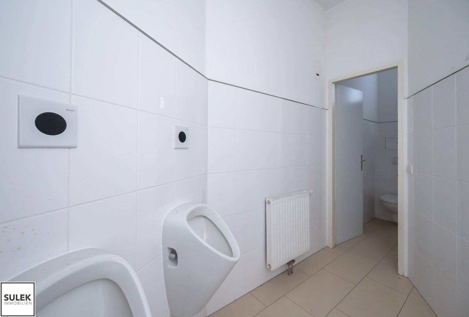 Sanitärraum mit Urinal, Toilette und Fliesen an Wänden und Boden, der renoviert werden muss.