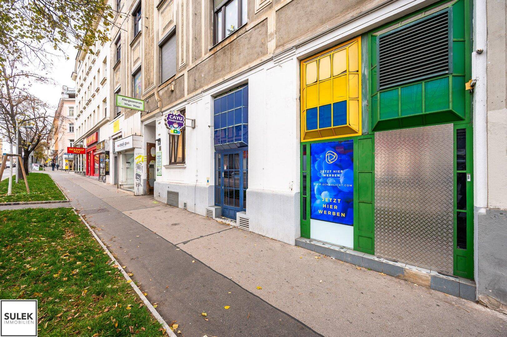 Erdgeschossfassade eines Altbaus mit bunten Schaufenstern und einem Bürgersteig in der Stadt.