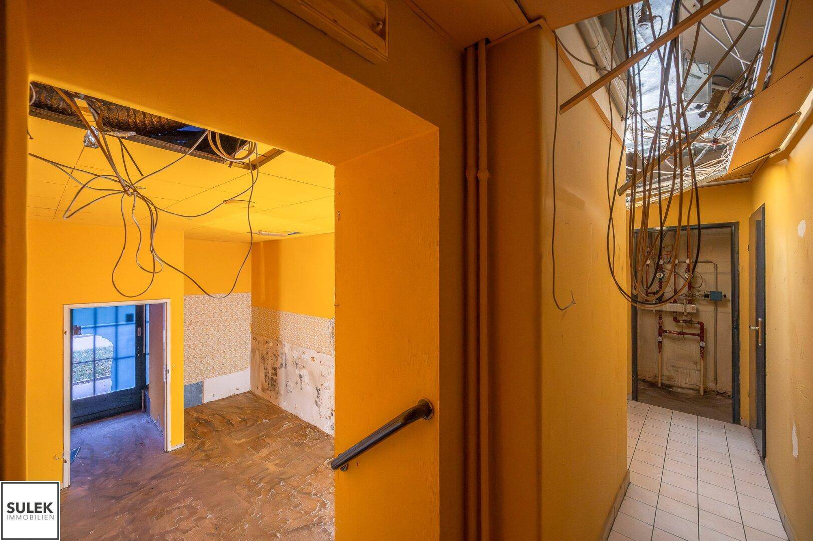 Flur in Renovierungszustand mit gelben Wänden, freiliegenden Kabeln und Rohren an Decke und Wand.