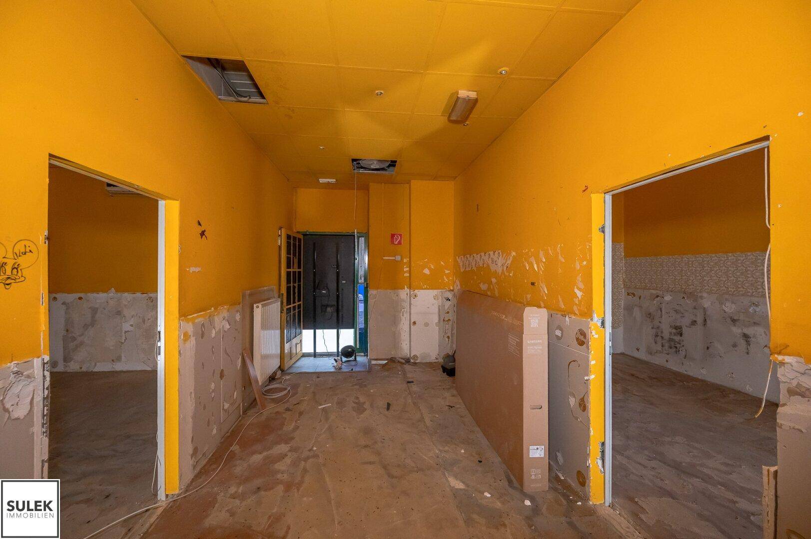 Großer Raum in Renovierungszustand mit gelben Wänden, freigelegten Wänden und mehreren Türöffnungen.