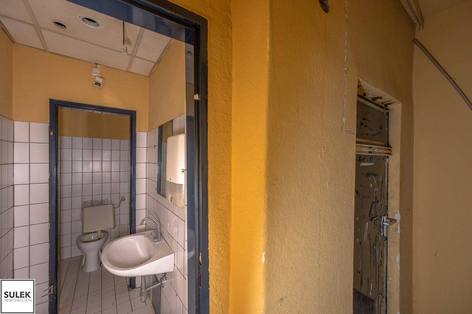 Einfacher Toilettenraum in Renovierungszustand mit weißen Fliesenwänden, WC und Waschbecken.