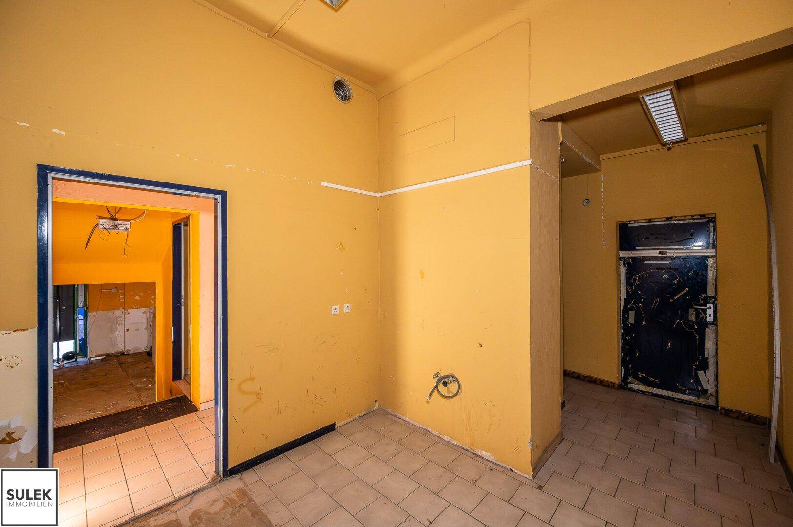 Flur in Renovierungszustand mit gelben Wänden, gefliestem Boden und Blick auf einen weiteren Raum.