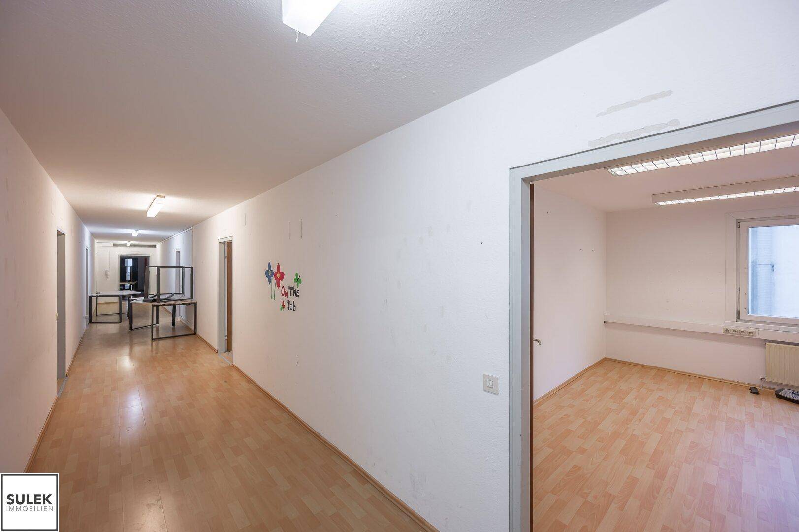 Langer Flur mit Holzfußboden und weißen Wänden, der zu verschiedenen Büroräumen führt.