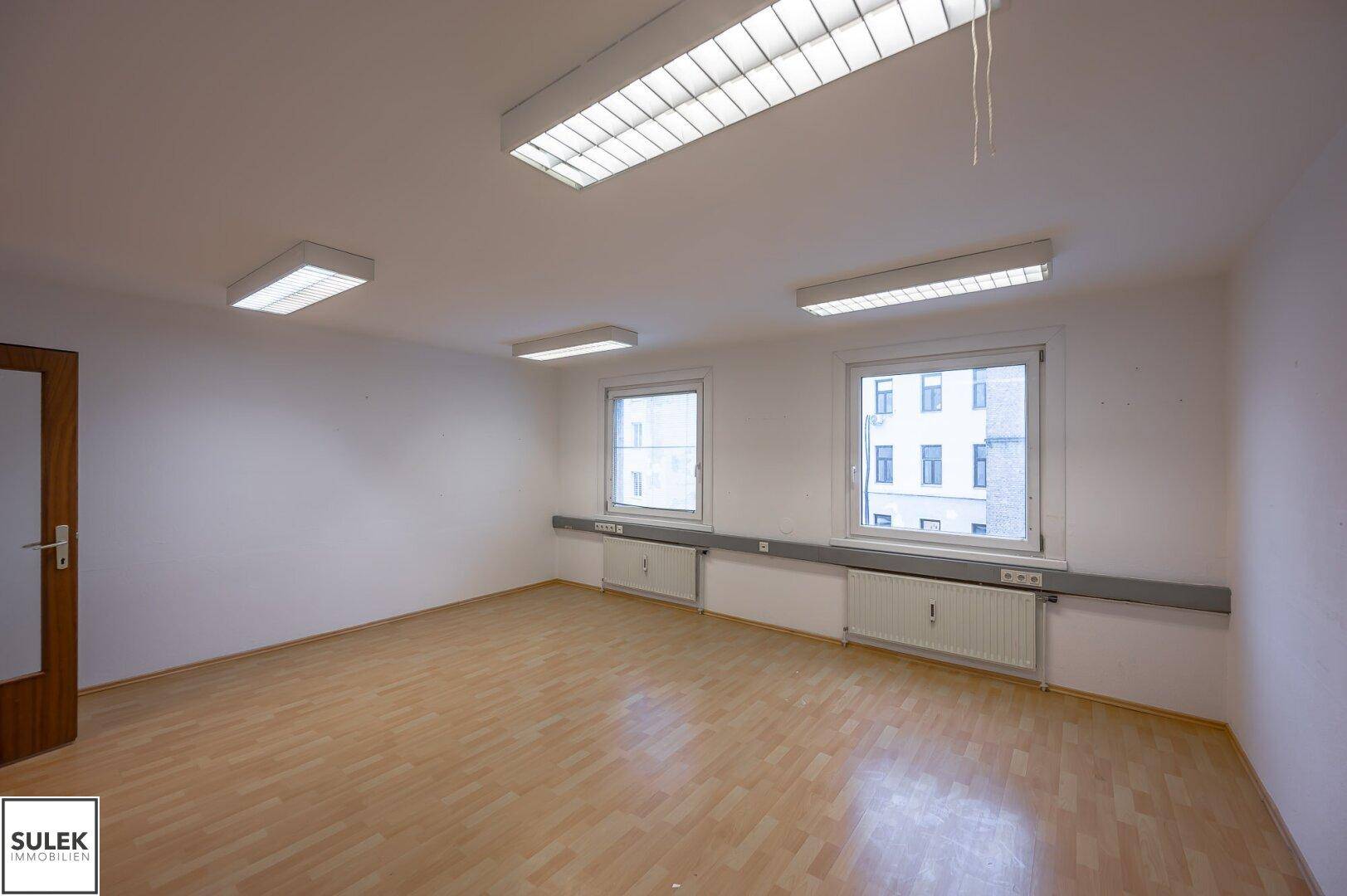 Heller, leerer Raum mit Holzfußboden und zwei Fenstern, die auf die Stadt blicken.