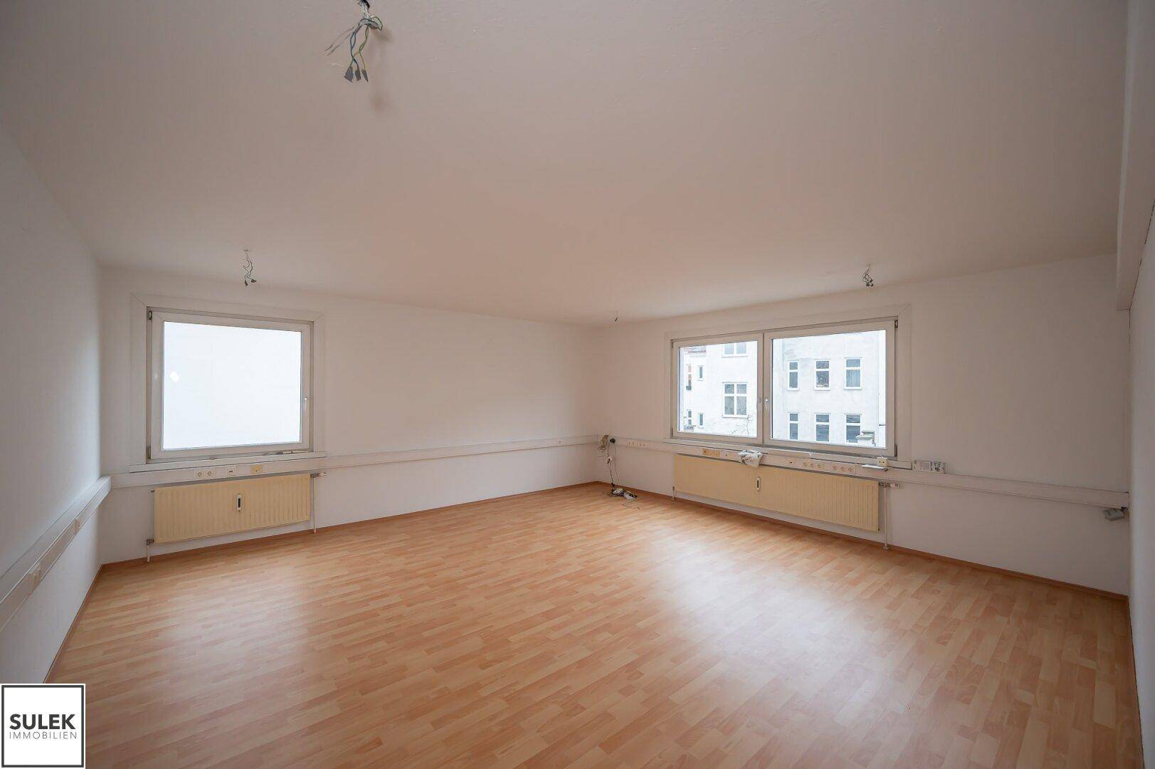 Leerer Raum mit Holzfußboden und zwei Fenstern, die eine helle Atmosphäre schaffen.