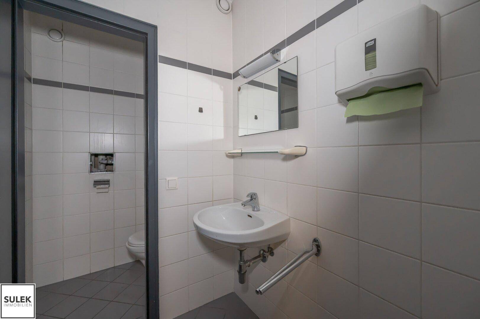 Einfache Toilette mit weißen Fliesen, Waschbecken und Haltegriffen für Barrierefreiheit.