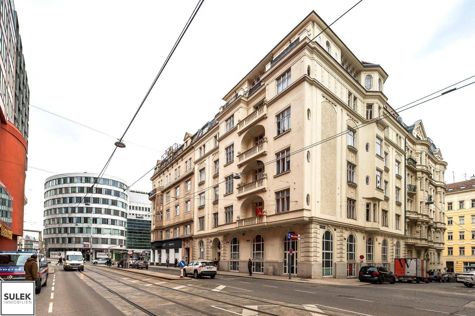 Historisches Eckgebäude an einer belebten Straße mit Straßenbahnlinien und modernen Gebäuden im Hintergrund.