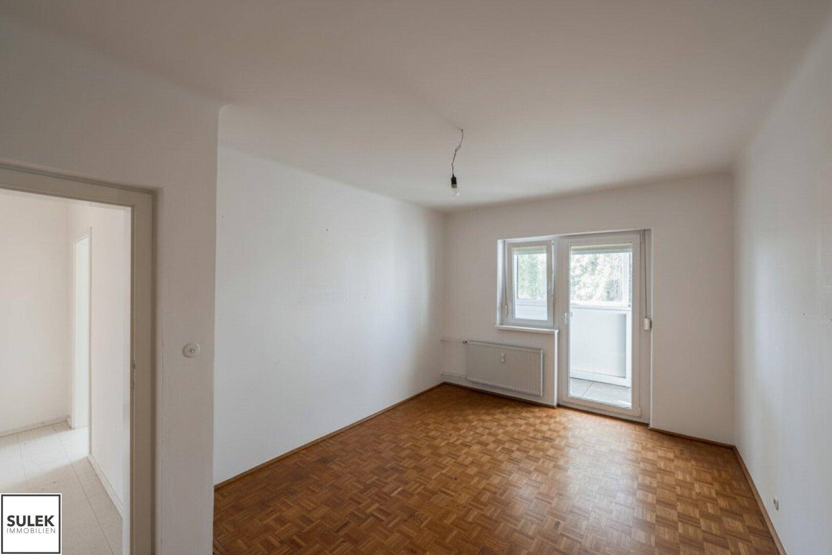 Unmöbliertes Zimmer mit Parkettboden und Fenster, das viel Tageslicht hereinlässt.