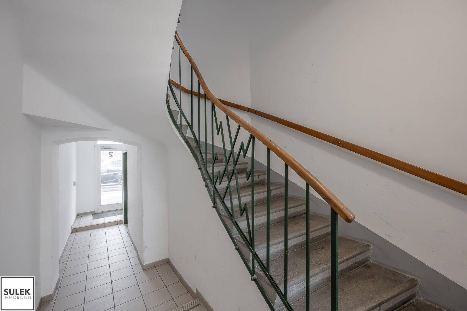 Treppenhaus mit Steinstufen, grünem Metallgeländer und Holzhandlauf, führt zu einem gefliesten Flur.