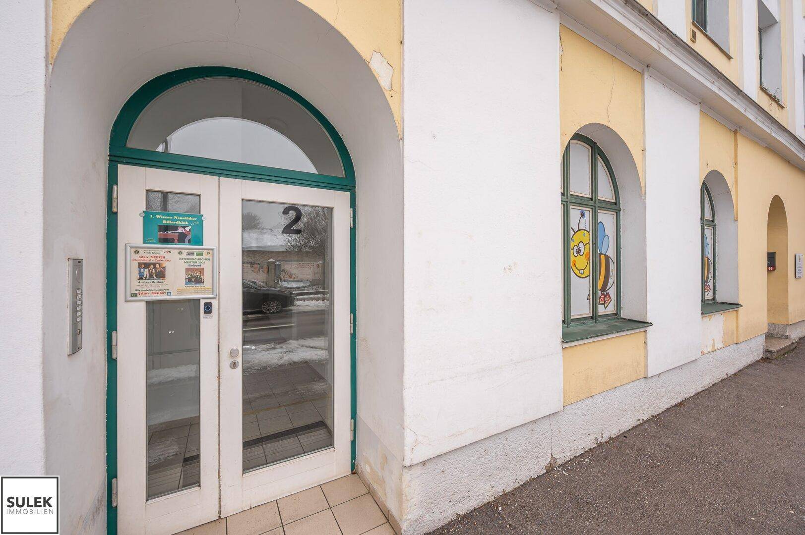 Eingangsbereich Nummer 2 mit grünen Türrahmen und einem Fenster mit Bienenmotiv.